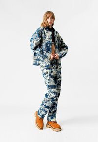 Conjunto de chaqueta y pantalones de mezclilla en un patrón abstracto en azul y blanco, con un corte relajado, acompañado de una blusa negra corta y botas de tobillo en color beige.