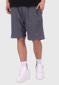 Graue gerippte Shorts mit einem elastischen Bund, ausgestattet mit Seitentaschen und einem kleinen Logo am unteren Bein, kombiniert mit weißen Sneakers.