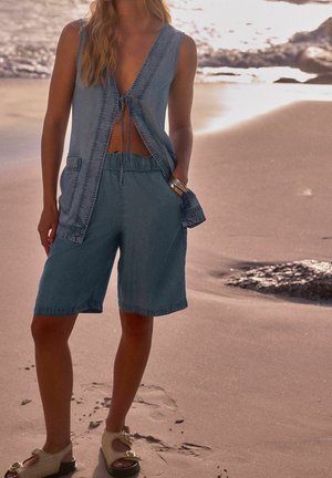 Chaleco de mezclilla azul claro con escote en V y lazo en la parte delantera, combinado con pantalones cortos holgados a juego y sandalias beige, en una playa de arena.