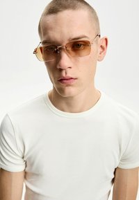 Hnedé obdĺžnikové slnečné okuliare s tenkými kovovými rámy a detailne spracovanými rámami. Nosený je obyčajný biely tričko s rebrovaním.