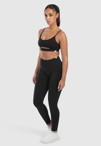 Zwarte sportieve crop top met een uitsnijding en dunne bandjes, gecombineerd met hoge zwarte leggings. Model draagt witte sneakers.