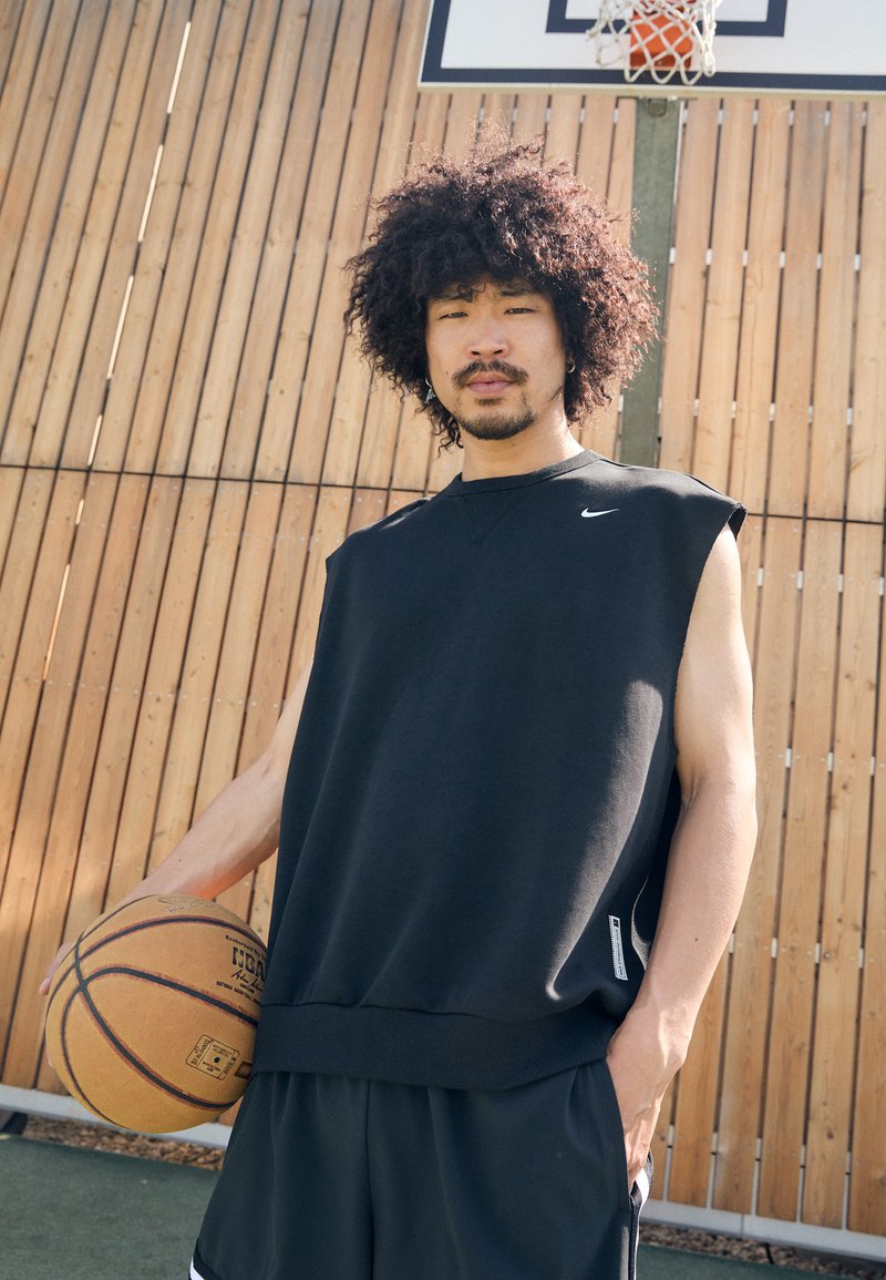 Svart ermeløs treningsdrakt med rund hals og ribbestrikket kant, kombinert med svarte shorts. Holder en brun basketball mot en trebakgrunn.