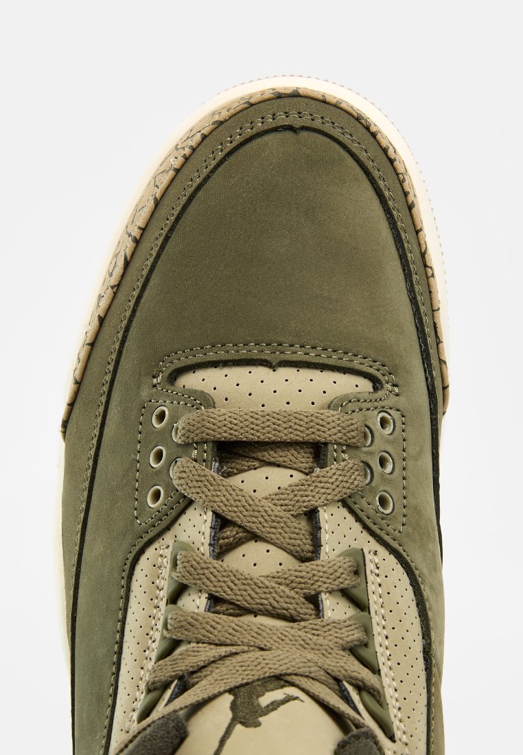 Gros plan d'une sneaker vert olive et beige avec des lacets texturés et des panneaux latéraux perforés sur un fond uni.