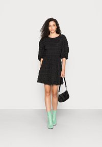 Vestido preto com mangas bufantes, com babados texturizados, cintura ajustada e saia evasê. Combinado com botins verdes-menta e uma pequena mala preta.