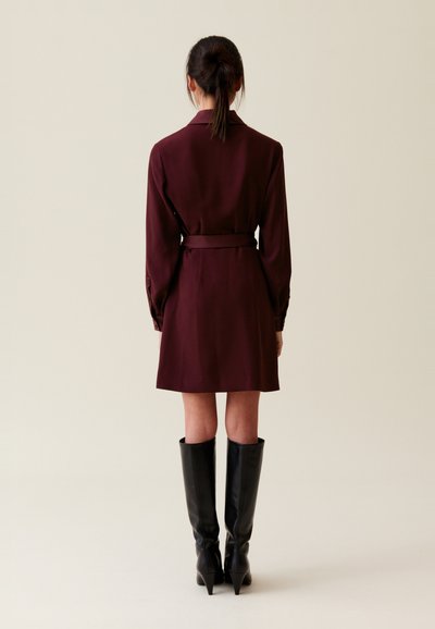 Vestido burdeos con mangas largas, cintura ceñida con cinturón y cuello. Combinado con botas negras hasta la rodilla. Vista trasera contra un fondo neutro.