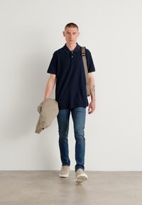 Námornícka polo košeľa, krátke rukávy, klasický golier; nesúca svetlo béžovú bundu; úzke džínsy; béžové tenisky; neutrálny pozadie.