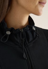 Černá bunda na zip s vysokým límcem, s žebrovanou texturou, kovovým zipem a nastavitelnými stahovacími šňůrkami na límci.