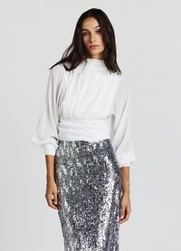 Blusa bianca plissettata con scollo alto e maniche ampie, abbinata a una gonna aderente con paillettes argentate e motivo strutturato.