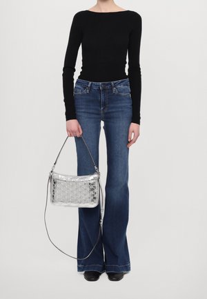 Frau trägt ein schwarzes langärmeliges Oberteil und blaue Jeans mit weitem Hosenbein und hält eine silberfarbene, geprägte Handtasche mit einem langen Riemen.