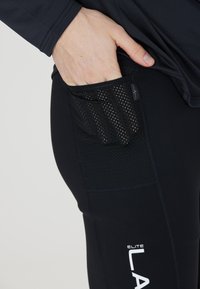 Black athletic leggings featuring a mesh pocket on the side. Smooth texture with a fitted design, branded with white text.