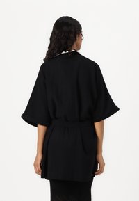 Veste de style kimono noire avec des manches larges, une taille cintrée et une texture lisse. Design simple sans motifs ni embellissements.