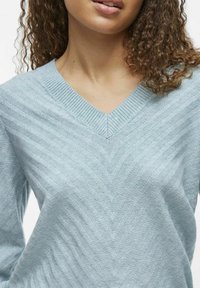 Lyseblå strikket genser med V-hals, med et strukturert chevronmønster og ribbestrikkede mansjetter. Mykt materiale og avslappet passform.