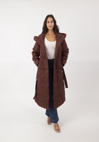 Abrigo largo acolchado marrón con capucha, que presenta una cintura ceñida con cinturón, textura acolchada y bolsillos laterales, combinado con jeans azules y sandalias.