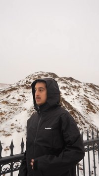 Man in black hooded jacket standing by a snow-covered rocky hill with an iron fence in front under overcast sky.