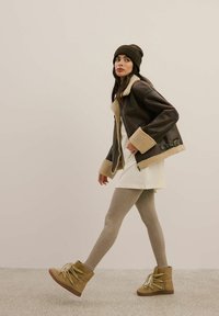 Brązowa skórzana kurtka z owczą wełną przy kołnierzu, biała sukienka, jasnobeżowe leginsy i beżowe buty na sznurówki; modelka pozuje na teksturowanej podłodze.