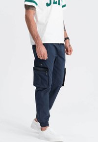 Pantalons cargo navy avec des poignets élastiques et de multiples poches latérales. Fabriqués en tissu durable, avec une texture lisse et des détails zippés.