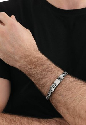 Pulsera trenzada en plata y negro con un cierre metálico suave, que presenta un diseño texturizado y un destacado detalle central.