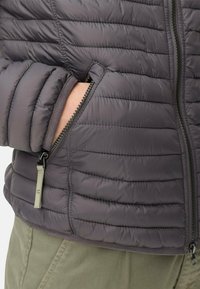 Grijze gewatteerde jas met een ritszak, horizontaal gestikt ontwerp en een gladde textuur. Geaccentueerd met een contrasterende rits.