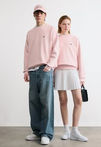 Νέος άνδρας και γυναίκα που φορούν αντιστοιχισμένα ανοιχτό ροζ φούτερ, λευκά αθλητικά παπούτσια· ο άνδρας φορά φαρδιά τζιν και καπέλο, η γυναίκα λευκή πλισέ φούστα και κρατάει μαύρη τσάντα.