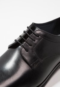 Zapato de vestir de cuero negro con una superficie suave, seis ojales para los cordones y un sutil detalle de costura a lo largo del lado.