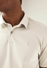 Camisa polo beige con cuello, tapeta de dos botones y un pequeño logo de ciervo bordado en el lado izquierdo del pecho. Textura de algodón suave.