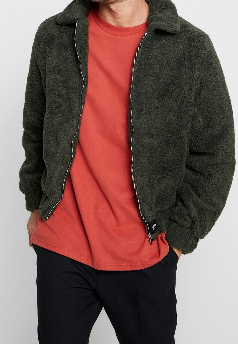 Olijfgroene, zachte fleecejas met een ritssluiting aan de voorkant en een kraag. Gecombineerd met een rood shirt en zwarte broek, wat een casual stijl toont.