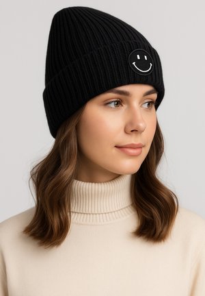 Black knitted beanie with ribbed texture and a white smiley face patch on the front. Worn with a beige high-neck sweater.