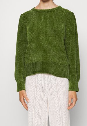 Personne portant un pull texturé vert olive à manches longues associé à un pantalon en dentelle blanche taille haute sur un fond uni.
