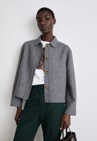 Casaco de lã cinza com gola, apresentando seis botões de latão. Usado com uma camisa branca e calças verdes. Textura suave.