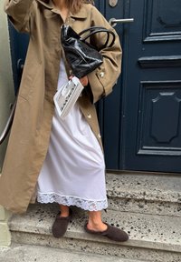 Vaaleanruskea trench-coat suurilla napeilla valkoisen mekon päällä, jossa on pitsireuna. Asusteena pörröiset ruskeat tossut ja musta teksturoidusta materiaalista valmistettu käsilaukku.