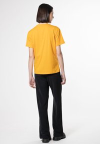 Gul bomulds t-shirt med korte ærmer og rund hals, parret med sorte vide bukser, vist fra ryggen mod en hvid baggrund.