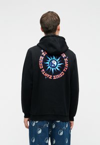 Fekete kapucnis pulóvert viselő személy, amelynek hátrészén kék nap és yin-yang szimbólum, valamint narancssárga kör alakú "Santa Cruz" felirat található, amelyet yin-yang mintás farmernadrággal párosítanak.