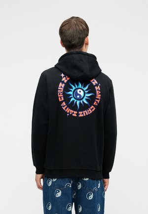 Henkilö, joka on pukeutunut mustaan huppariin, jossa on sininen aurinko ja yin-yang symboli sekä oranssi pyöreä teksti "Santa Cruz" selässä, yhdistettynä yin-yang -kuvioisiin farkkuihin.