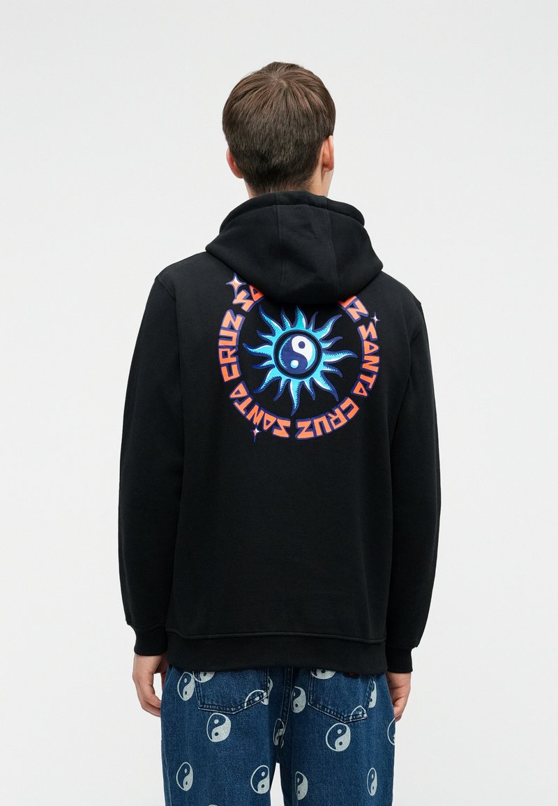 Fekete kapucnis pulóvert viselő személy, amelynek hátrészén kék nap és yin-yang szimbólum, valamint narancssárga kör alakú "Santa Cruz" felirat található, amelyet yin-yang mintás farmernadrággal párosítanak.