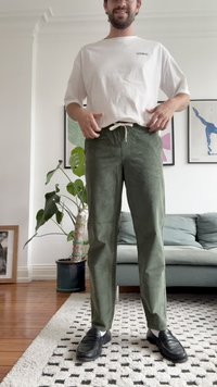 Helles T-Shirt mit einem Logo, grüne Hose mit Kordelzug, und schwarze Schuhe, vor Zimmerpflanzen und gerahmter Kunst.