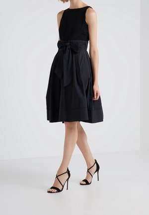 Schwarzes ärmelloses Kleid mit einem taillierten Oberteil und einem schwingenden Rock, der mit einer großen Schleife in der Taille verziert ist und eine glatte Textur aufweist. Kombiniert mit schwarzen Riemchensandalen.