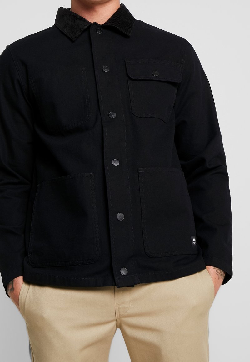 Svart jacka med en kordflätskrage, med två bröstfickor och två stora framfickor, knäppning med knappar och ett något texturerat tyg.