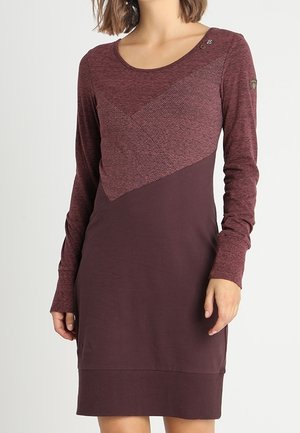 Vrouw draagt een knielange jurk met lange mouwen in twee tinten bordeauxrood met een gestructureerd diagonaal paneel over de borst en schouder.