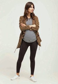 Casaco castanho leve, topo às riscas preto e branco, leggings pretas e ténis brancos. Modelo com os braços cruzados.