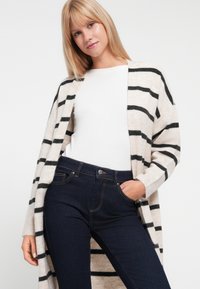 Beige und schwarz gestreifter Cardigan aus weichem Stoff, mit offenem Vorderteil, kombiniert mit dunkelblauen Jeans im klassischen Fünf-Taschen-Stil.