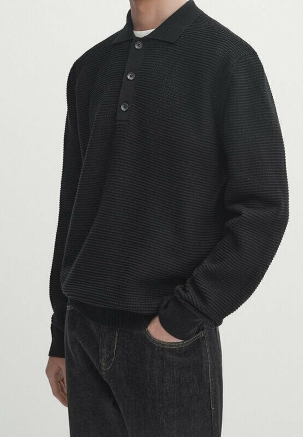 Pull noir texturé avec un col polo, trois boutons et des poignets côtelés. Associé à un jean sombre, affichant un style décontracté.