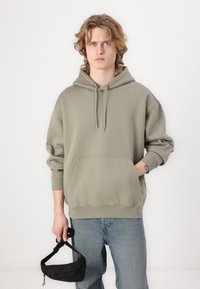 Olive green hooded sweatshirt made of soft fleece material, featuring a front pocket, relaxed fit, and ribbed cuffs, paired with denim jeans.
