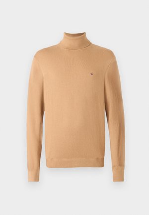 Beige højhalset sweater med lange ærmer, glat tekstur og en lille logoaccent på venstre bryst. Letvægts- og tætsiddende design.