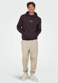 Haut à capuche violet foncé avec poche kangourou et texte blanc, associé à un pantalon de survêtement beige et des baskets de couleur claire. Design à coupe décontractée.