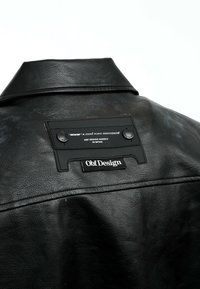 Chaqueta de cuero negro con cuello abotonado, una etiqueta de tela en la parte posterior y un acabado texturizado. La marca destacada incluye "Obf Design."