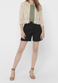 Cardigan corto in pizzo crema sopra una maglietta verde, abbinato a pantaloni corti neri e sandali con tacco marroni. Presenta motivi in pizzo intricati.