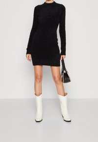 Kobieta stojąca w czarnej, puszystej mini sukience z długim rękawem, trzymająca czarno-białą torebkę, nosząca białe kowbojki do połowy łydki.