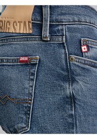 Közeli kép kék farmernadrágról, dupla hátsó zsebekkel, narancssárga varrással, piros márkajelzéssel és bézs bőrcímkével a derékszíjon.