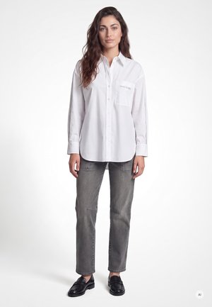 Woman with long wavy hair wearing a white button-up shirt and gray jeans standing against a plain white background.