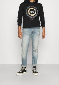 Sudadera negra con estampado gráfico, pantalones vaqueros de mezclilla azul claro y zapatillas altas negras, con cordones blancos y un detalle de dobladillo enrollado.